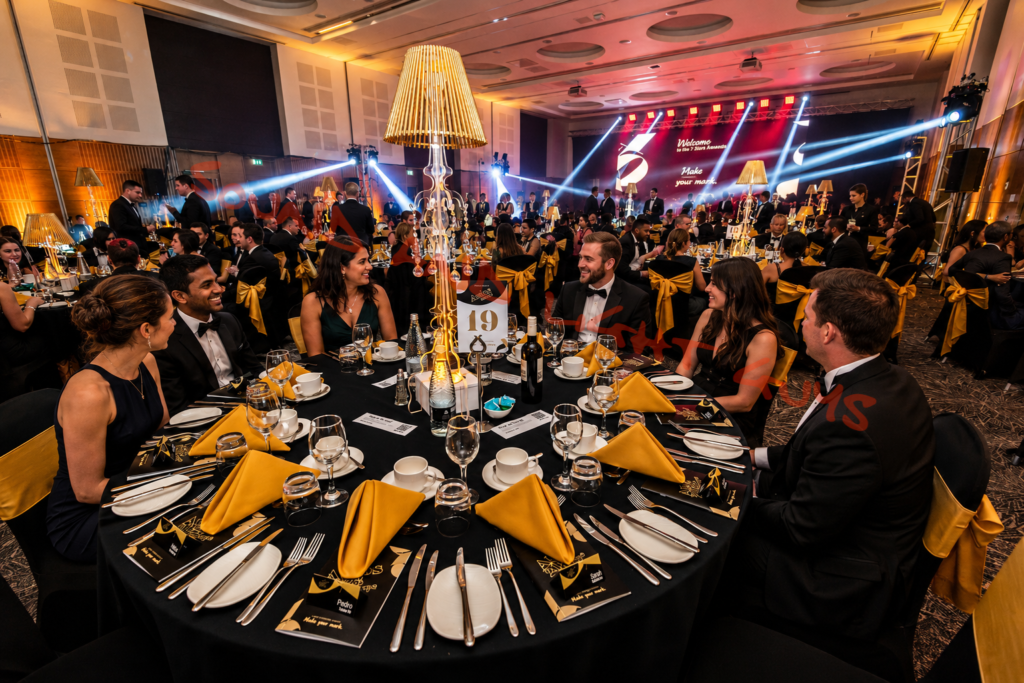 Professional corporate event production setup with stage lighting, banquet tables, AV equipment, and guests at a luxury gala event.
